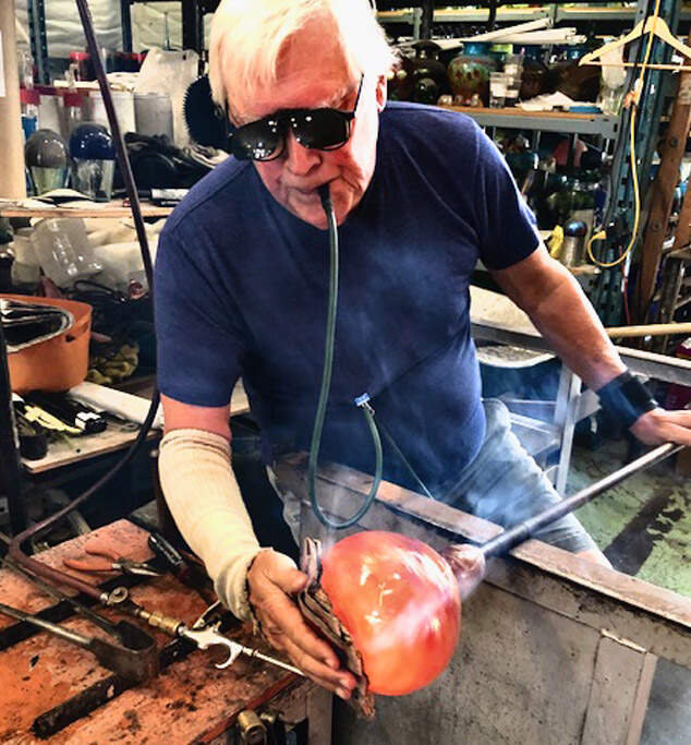 Robert Held using tube to blow into heated glass piece with sunglasses on